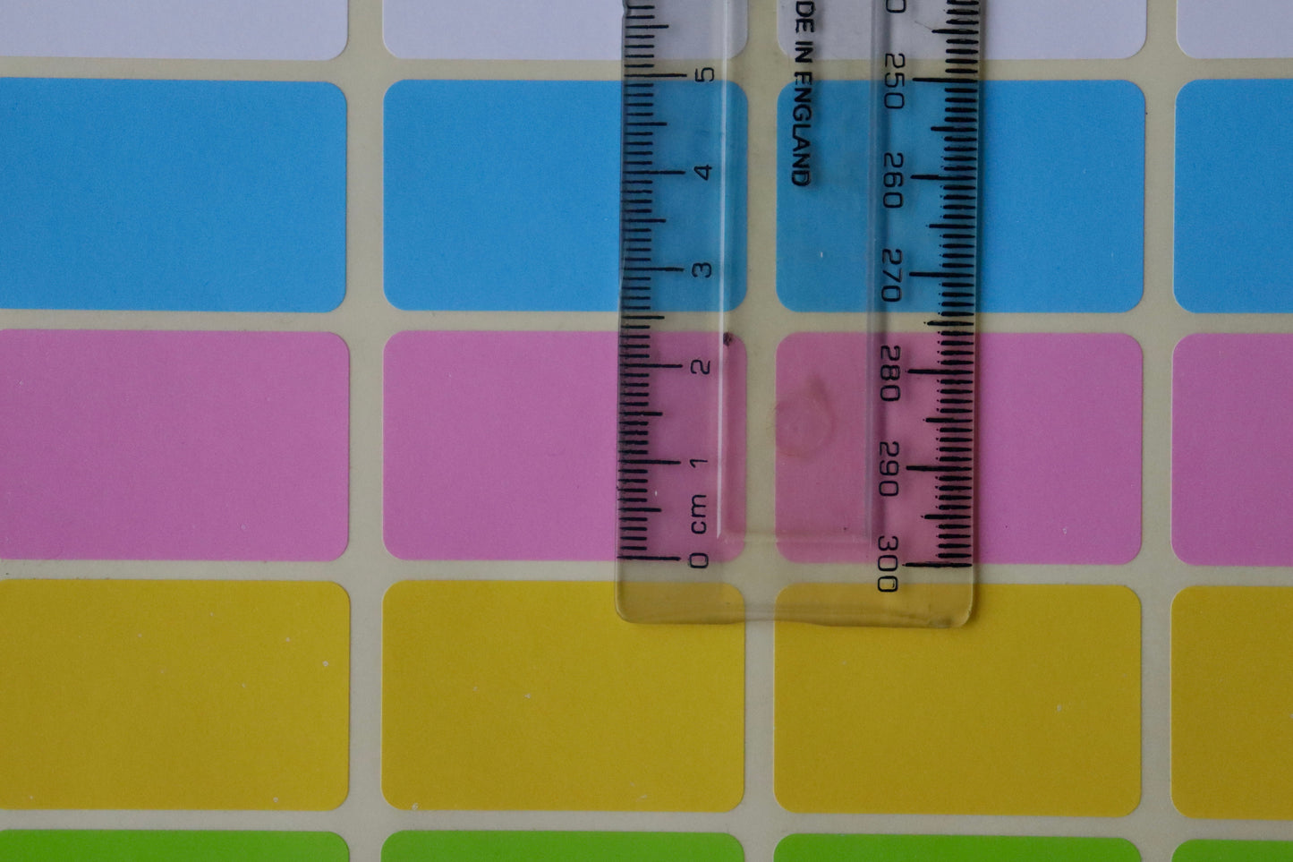 A clear plastic ruler measuring blue, pink, and yellow rectangular stickers on a sheet.
