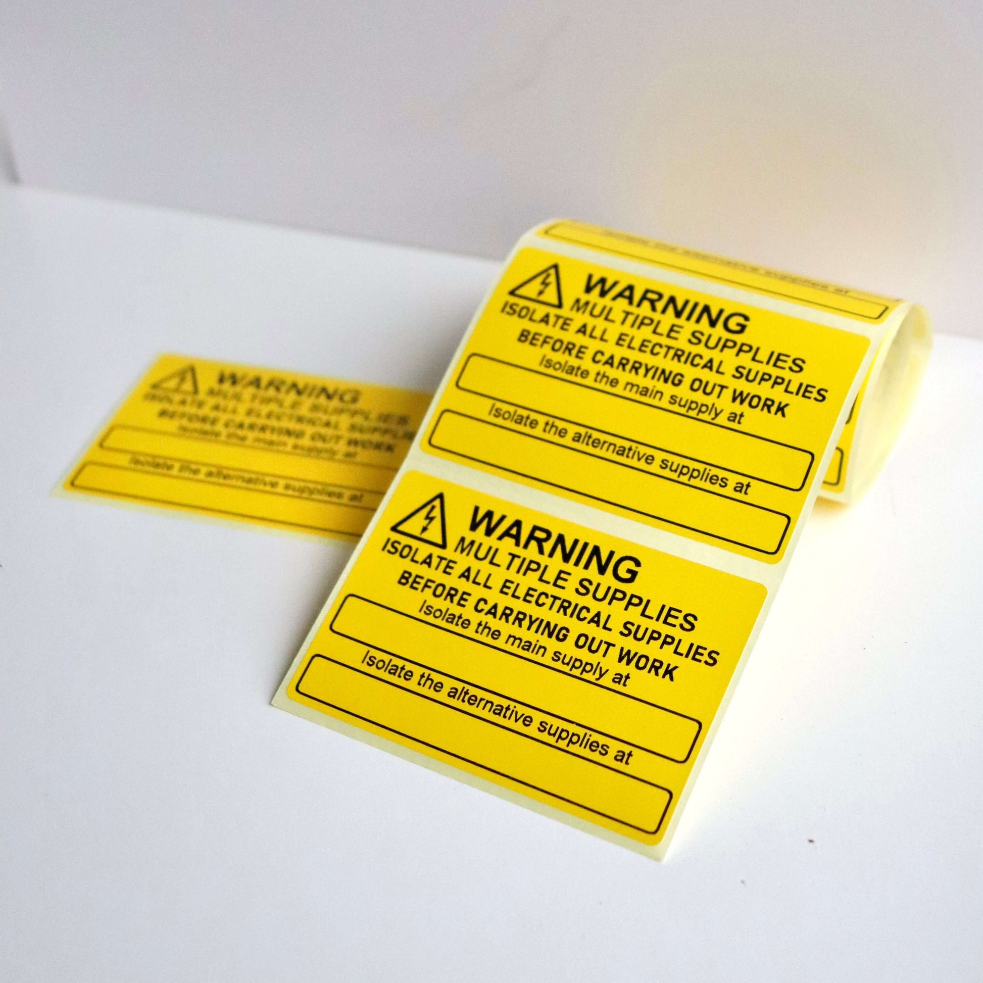 Yellow warning labels with black text and exclamation icons, rolled and unrolled on a white surface.